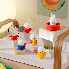 Six small white bunny figurines in colorful clothes playing with various toys on white tiered platform