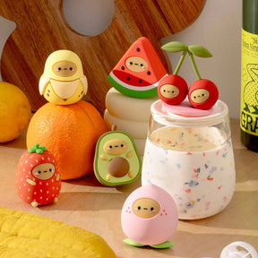 The Smoko Tayto Fruit Blind Box by Smoko features fruit-shaped silicone figures with faces, arranged next to a speckled jar, citrus fruits, and a bottle of olive oil.