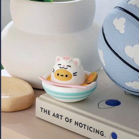 A small, collectible Smoko Tayto Potato Cat figurine sits in a striped bowl on a book titled "The Art of Nothing," near a vase and a blue ball with white clouds. It's like discovering a treasure from the Smoko Tayto Potato Cat Blind Box, reminiscent of the joy when you uncover the next adorable Tayto Potato!