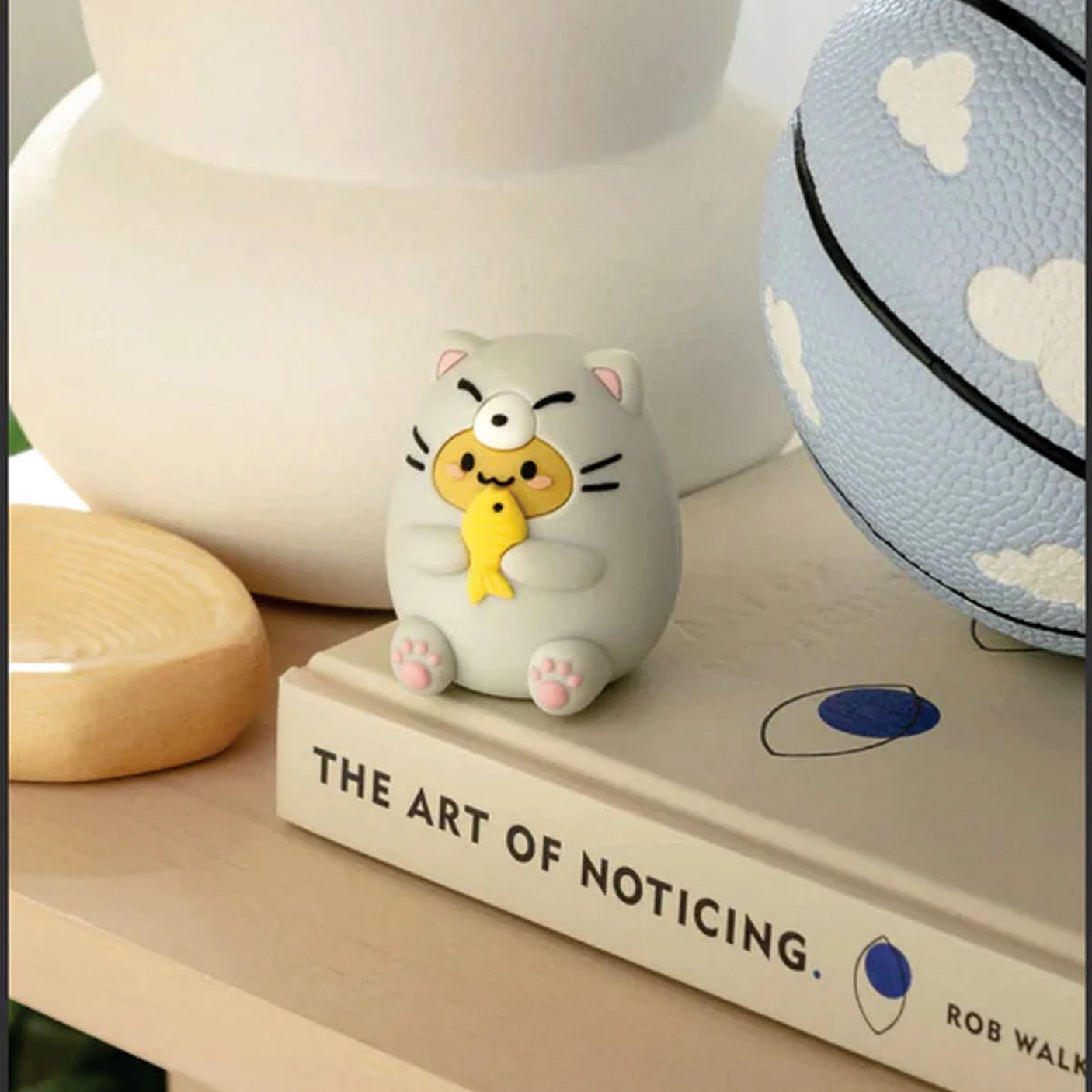 A Smoko Tayto Potato Cat Blind Box figurine, depicting a small cat holding a yellow fish, is perched on a book titled "The Art of Noticing." Nearby, the charming presence of a blue basketball adorned with clouds adds to its allure, evoking the delightful surprise of opening a Blind Box filled with enchanting collectibles.