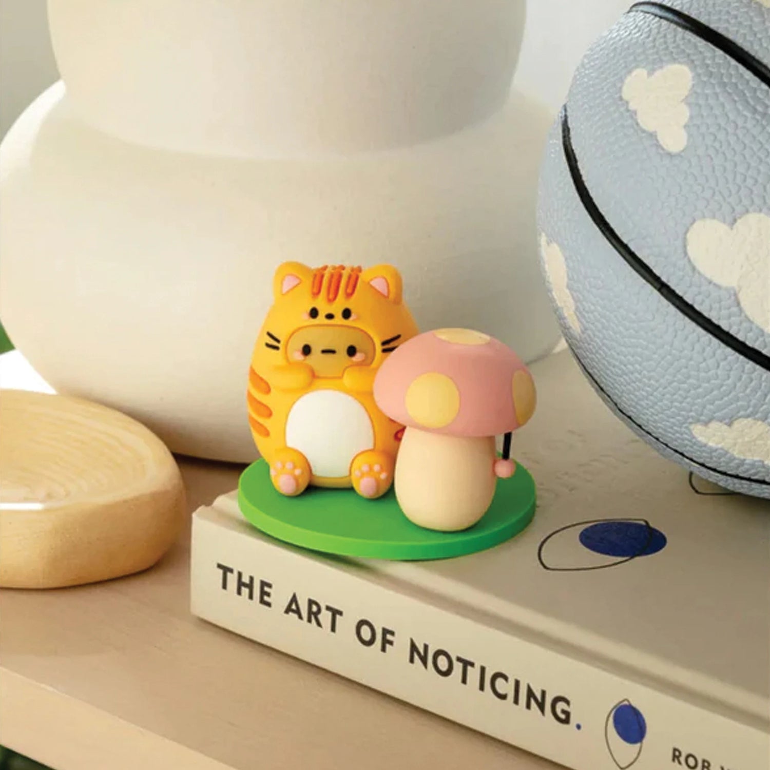 A small tiger-shaped figure with a mushroom relaxes on "The Art of Noticing," next to a painted basketball, as if hidden in a Smoko Tayto Cat Blind Box.