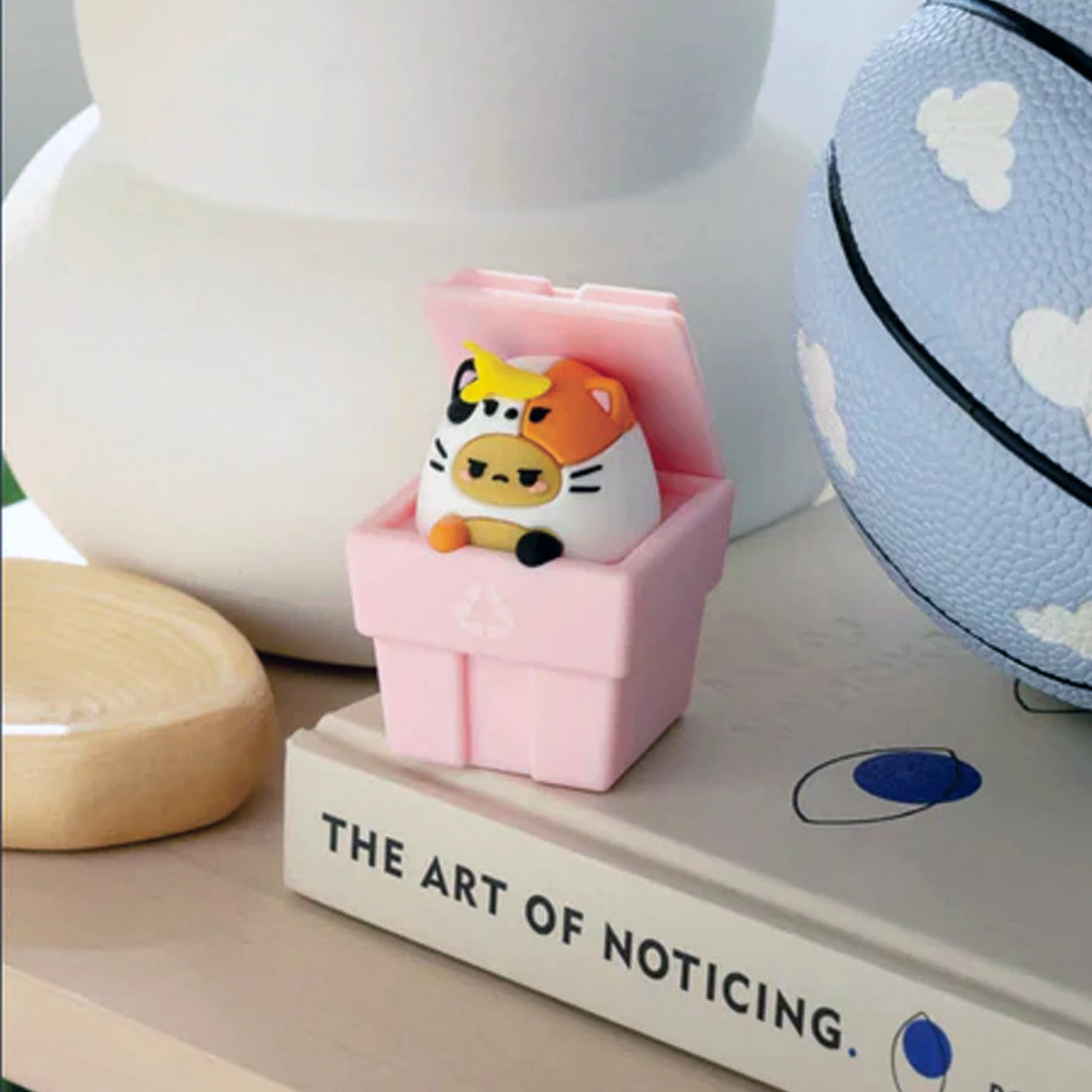 A Smoko Tayto Potato Cat Blind Box collectible figurine and a cow sit in a pink recycling bin on a book titled "The Art of Noticing," next to a basketball with cloud designs.