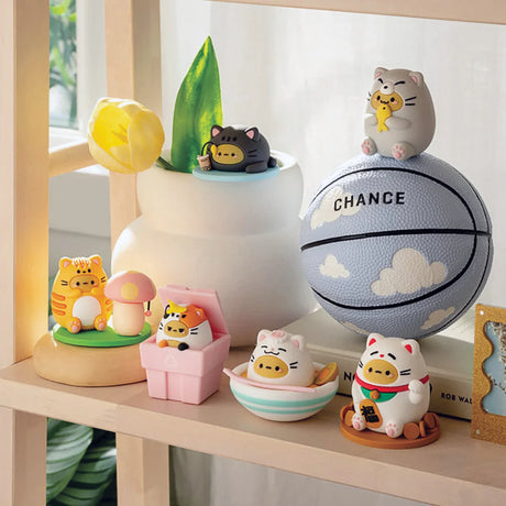 A shelf displays cat figurines in various poses and costumes, featuring the charming Smoko Tayto Cat Blind Box by Smoko, alongside a decorative basketball and a vase with a yellow tulip.