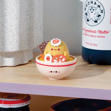 A Smoko Tayto Potato Ramen Blind Box vinyl figure featuring Gudetama lounging in a bowl can be seen on a wooden shelf, surrounded by bottles and other kitchen items.