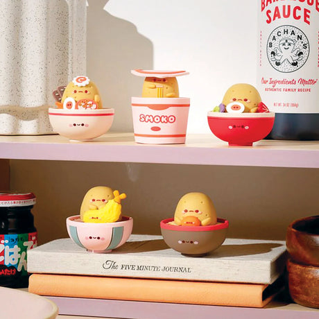 Cute yellow figures resembling dumplings from the Smoko Tayto Ramen Blind Box series are seated in bowls on two shelves, surrounded by sauces, a journal, and other items on a pink shelf.