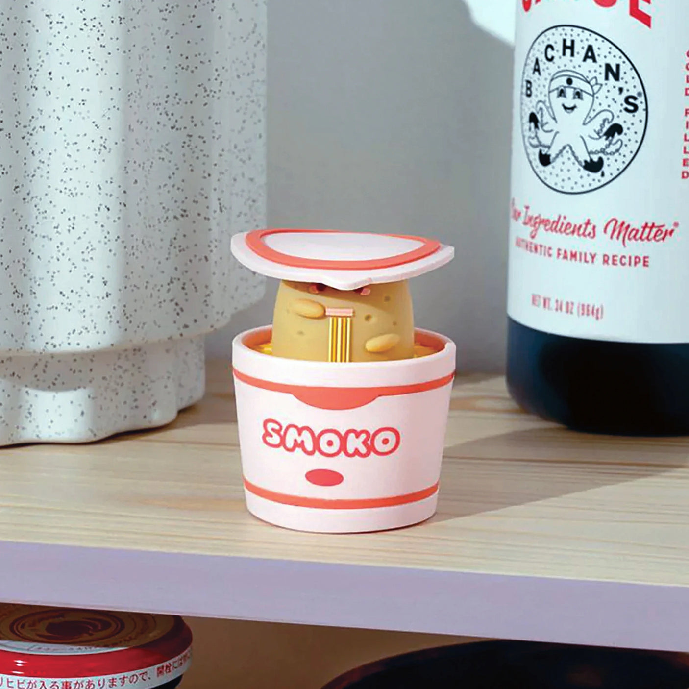 A delightful surprise, the Smoko Tayto Potato Ramen Blind Box vinyl figure emerges from a noodle cup container marked "Smoko," sitting on a wooden shelf next to a speckled container and a sauce bottle.