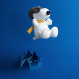 The Snoopy Plush Flying Ace - 18cm by the brand Snoopy is shown soaring over a blue paper pop-up design while wearing goggles and a scarf, set against a blue background.