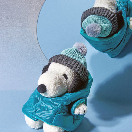 A Snoopy Plush in Puffer Jacket 20cm - Limited Edition from the Snoopy brand, dressed in a teal jacket and blue pom-pom beanie, sits before a circular mirror reflecting its upper half.