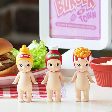 Three Sonny Angel Snack Series - Blind Box mini-figures by Sonny Angel stand before a burger and fries, each wearing a snack-themed hat like mustard, ketchup, or French fries. A food plate and sign are visible in the background.