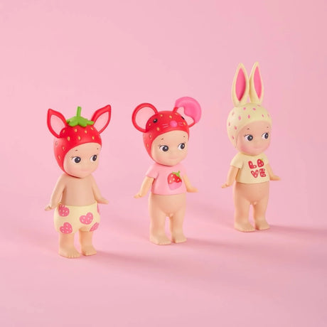 Three small dolls wearing fruit and animal-themed headpieces and pastel-colored clothing on pink background