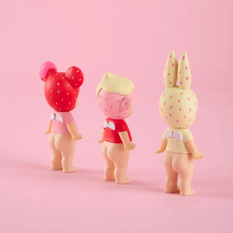 Three small doll figurines with strawberry-patterned headwear and wings on their backs facing away on pink background