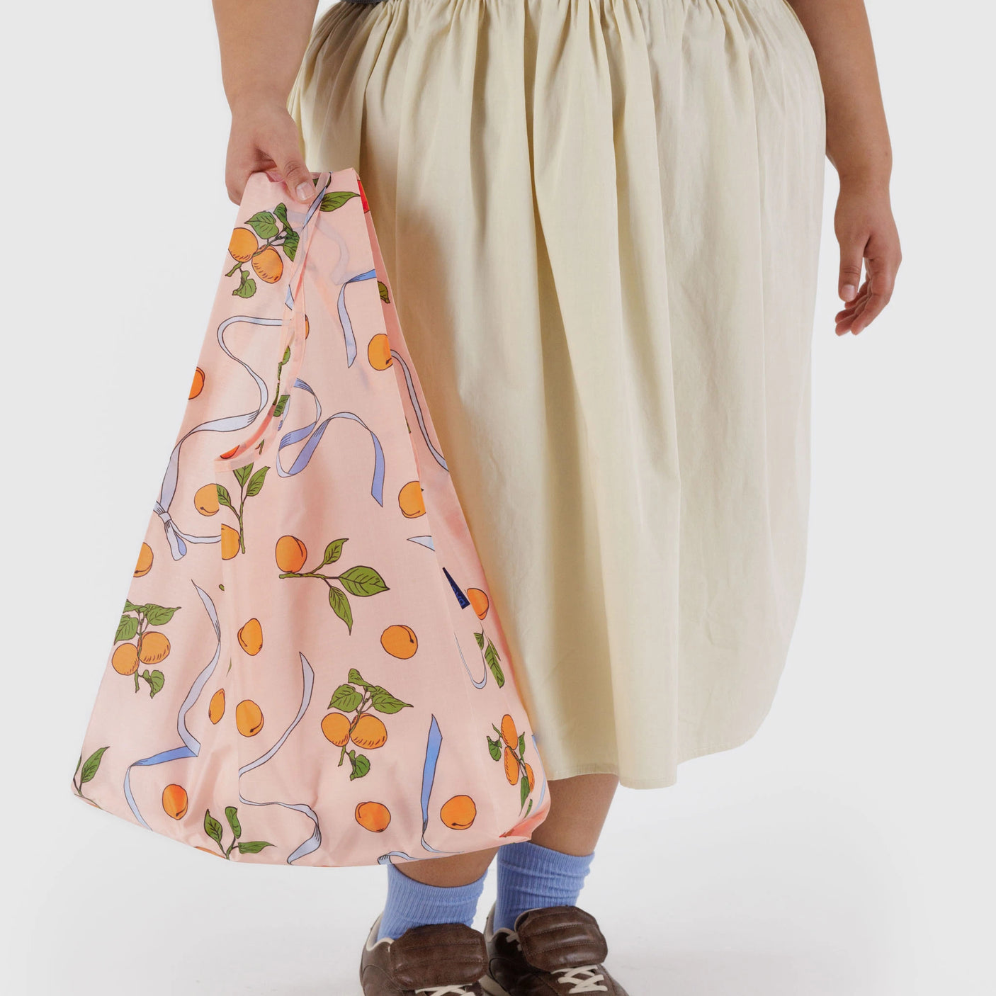 Someone is holding a Baggu Standard Baggu Bag - Apricots & Ribbons, standing against a white background in a cream skirt, brown shoes, and blue socks.