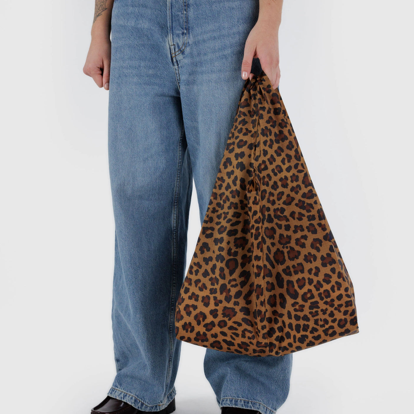 Against a plain background, someone holds a Standard Baggu Bag - Leopard by Baggu, made from recycled ripstop nylon. Their look is paired with blue jeans and black shoes.