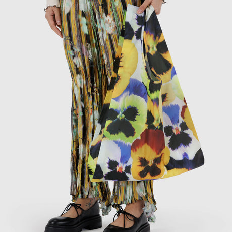 A person holds a vibrant Standard Baggu x Julia Heuer - Pansy shopping bag, crafted from recycled nylon, while wearing a pleated multicolored skirt and black shoes.