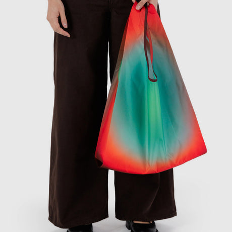 Someone carrying a Standard Baggu x Julia Heuer - Rain tote, featuring a stunning gradient of red, green, and blue, made from recycled nylon. They pair it with dark wide-leg pants and sleek black shoes.