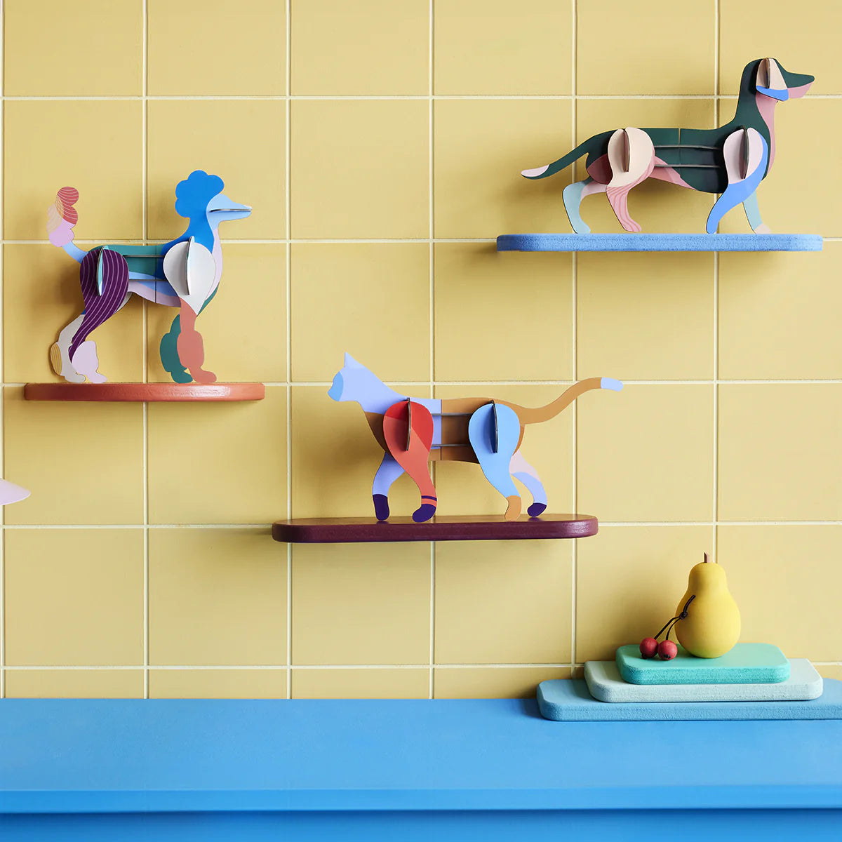 Studio Roof’s Wall Art Puzzle - Mochi Cat is showcased as playful wall art alongside colorful animal figurines, displayed on shelves with a yellow tiled background and fruit on a blue surface below.