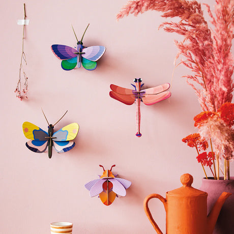 Studio Roof's Wall Art Puzzle - Blue Copper Butterfly is displayed with other vibrant paper insects as 3D wall sculptures, mounted on a pink wall near orange dried flowers and an orange teapot.