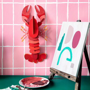 3D red lobster wall art puzzle mounted on pink grid wall with abstract painting on easel nearby gallery position 1