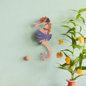Multicolored pink seahorse wall art puzzle mounted on pale green wall beside orange flowering plant gallery position 1