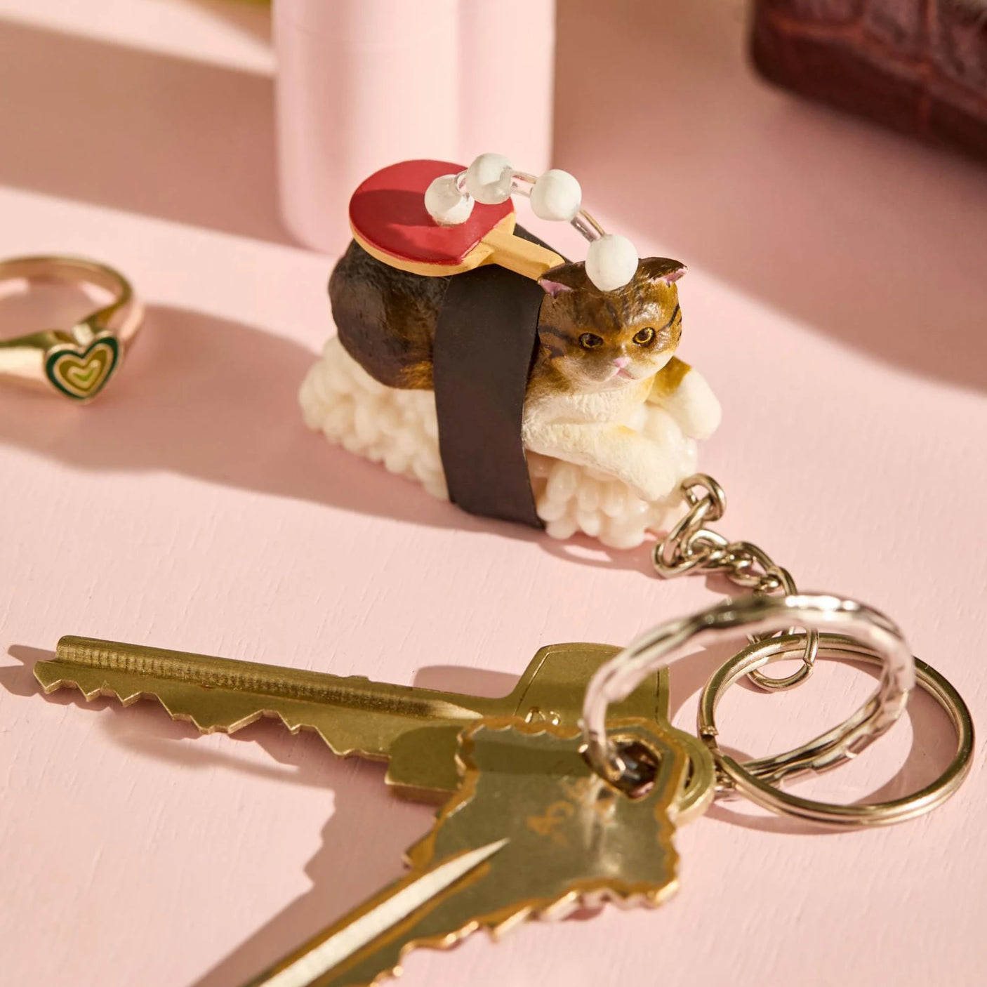 The Mofusand Sushi Cat Keychain Blind Box - Vol.2 figurine on a sushi-shaped keychain with a metal ring is artfully placed next to two brass keys, one with a heart-shaped ring, on a light pink surface.