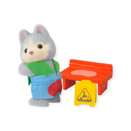 The Sylvanian Families Blind Bag - Baby Builder features a toy wolf in blue pants and a green scarf, standing by a red bench and yellow sandcastle caution sign—ideal for fans of Sylvanian Families collectibles.