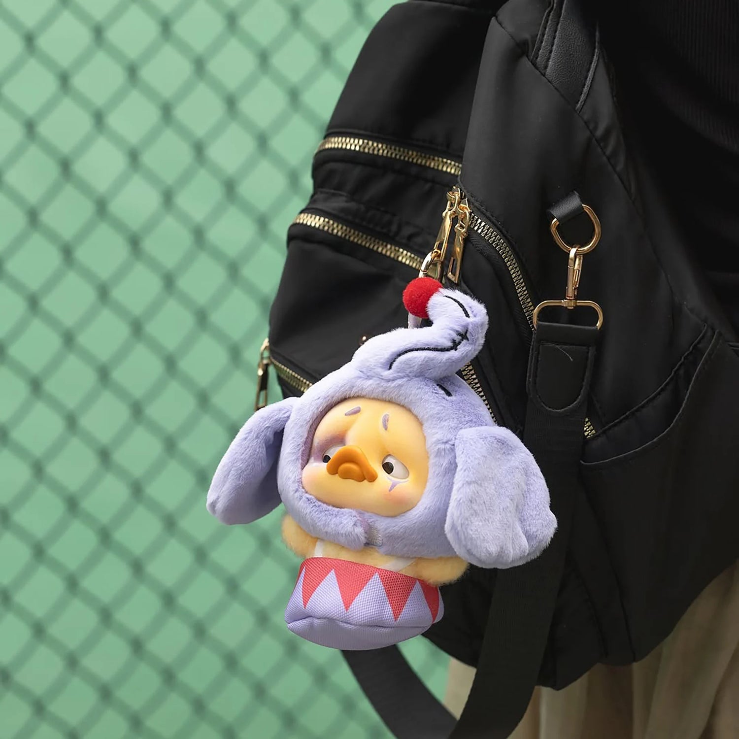 Lavender plush duck with bunny ears and red-and-blue striped outfit on a black backpack clip