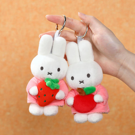 Two white plush bunny keychains in pink outfits holding a red strawberry and a red apple on orange background