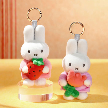 Two white plush bunny keychains dressed in pink holding a strawberry and a pink flower on yellow surface