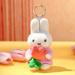 Plush white bunny keychain in pink outfit holding a red flower with green stem on yellow surface with petals nearby