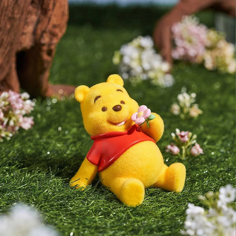 Yellow bear figurine in red shirt sitting on grass holding a small pink flower with blurred flowers in background
