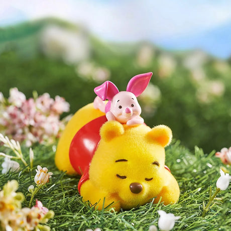 Small yellow bear figurine lying on grass with a pink-eared piglet resting on its back among flowers outdoors