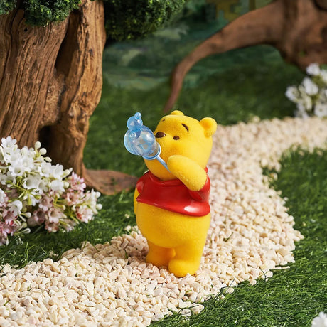 Small yellow bear figurine in red shirt blowing a blue bubble on a pebble path near tree and flowers outdoors