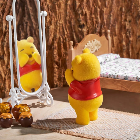 Yellow bear figurine in red shirt looking at itself in a standing white-framed mirror by a wooden bed with floral bedding