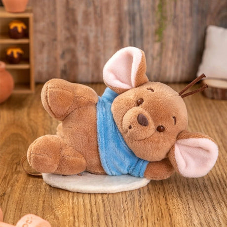 Plush brown mouse with large pink ears wearing a blue scarf lying on its side on a wooden floor