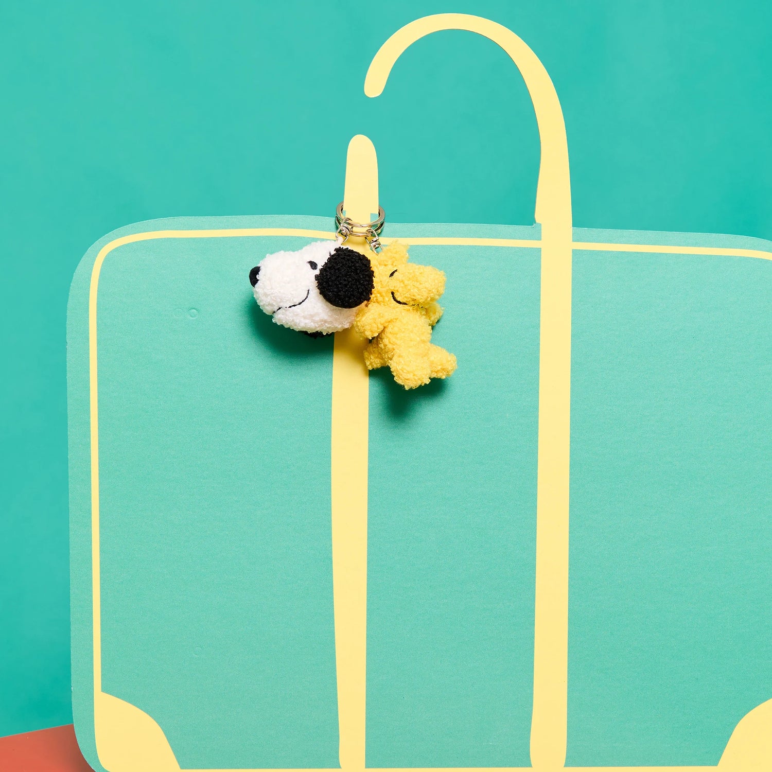 A Snoopy Woodstock Keychain 7cm by Snoopy is attached to the handle of a turquoise suitcase, shown against a teal background.
