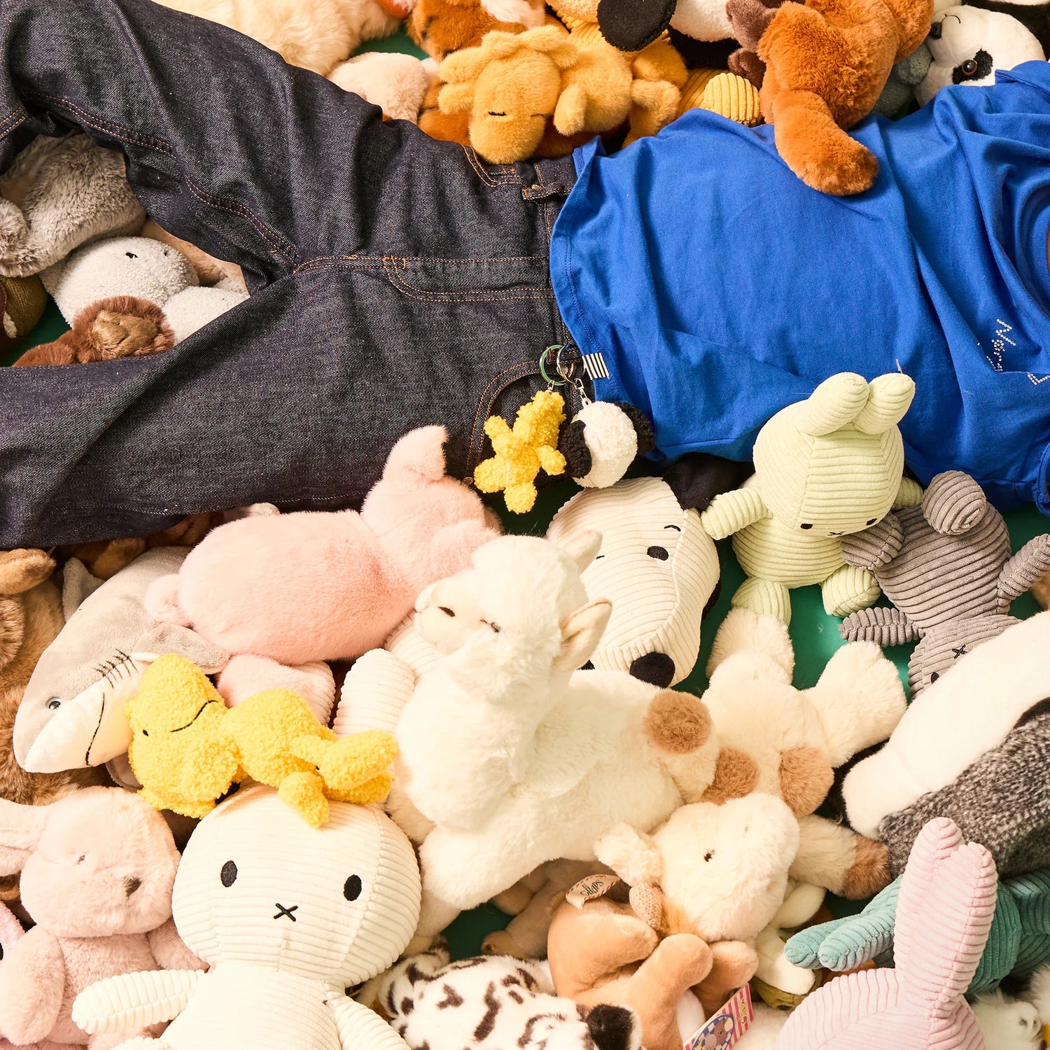 A person in a blue shirt and jeans lies on a large pile of assorted stuffed animals, plush toys, and a Snoopy Woodstock Keychain 7cm by Snoopy.
