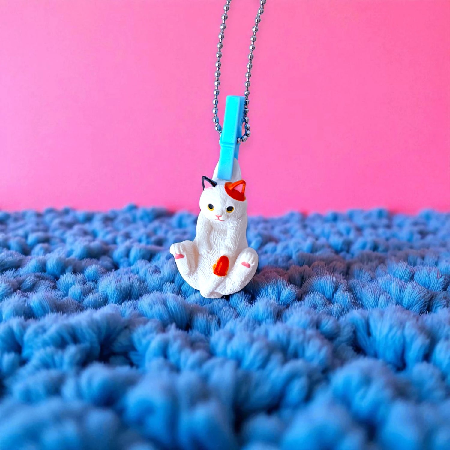 A Yell Washing Line Critters Charm Blind Box features a small white cat minifigure with orange and black spots on a textured blue surface, attached to a metal chain with blue clasp, set against a pink background.