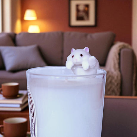 A small white hamster from Yell’s Wobble Cup Hamster Blind Box perches on a glass of milk in a cozy living room—an adorable desk toy for fans of cute collectibles.